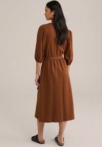 Brun, strukturerad midiklänning med puffärmar och bälte i midjan. Styld med svarta platta sandaler, visad bakifrån mot en neutral bakgrund.