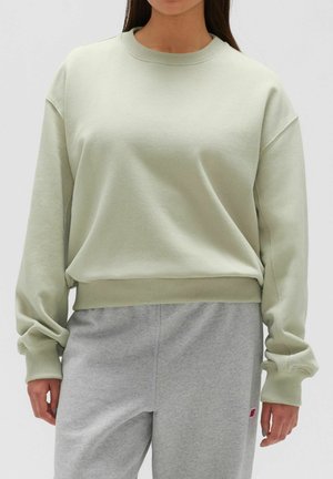 Femme portant un sweat-shirt oversize vert clair et un pantalon de jogging gris avec un petit logo rouge sur la cuisse gauche.