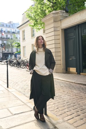 Femme portant un trench-coat sombre et un pull crème, debout les mains dans les poches dans une rue pavée près de vélos et d'un bâtiment beige.
