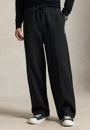 Personne portant un pantalon noir à jambes larges avec une taille à cordon, un haut noir à manches longues, des baskets noir et blanc et une montre-bracelet.