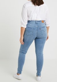 Jeans de tiro alto en azul claro con un ajuste slim, fabricados en denim. Cuentan con bolsillos traseros y un parche de marca de cuero. Llevados con una camisa blanca.