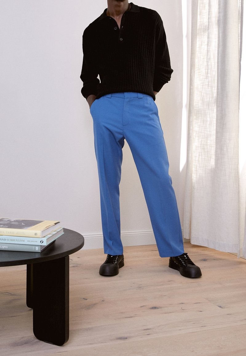 Uomo con maglione nero e pantaloni azzurri vivaci in piedi accanto a un tavolo nero con libri impilati in una stanza ben illuminata.