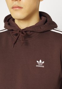 Sudadera marrón de algodón con cuello con cordón, que presenta tres rayas blancas en las mangas y un logo de Adidas bordado en blanco.
