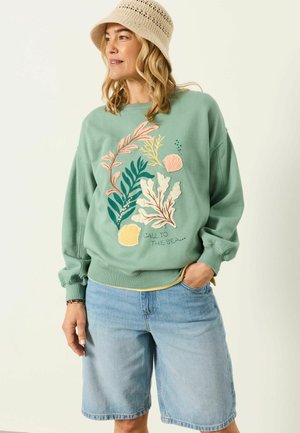 Woman wearing a beige crochet bucket hat, green sweatshirt with seaweed and shells design, and light blue knee-length denim shorts.