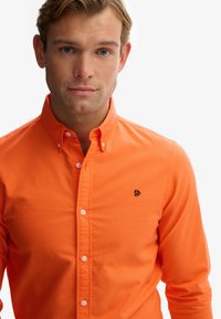 Młody mężczyzna o niebieskich oczach, ubrany w jasnopomarańczową koszulę na guziki z małym haftowanym logo na lewej piersi, stojący na białym tle.