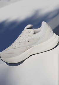 Baskets de sport blanches avec panneaux en mesh et en suède, semelle épaisse et rembourrée, et lacets, placées sur une surface blanche avec un fond ombragé.
