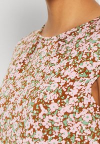 Blouse marron à motif floral avec des fleurs roses et vertes, dotée de manches courtes et d'un pli subtil au niveau de l'encolure. Texture de tissu douce.