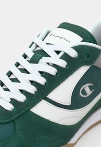 Adidași din suede verde cu accente din piele albă, având șireturi albe, un guler căptușit și o talpă din cauciuc. Logo brodat pe lateral.