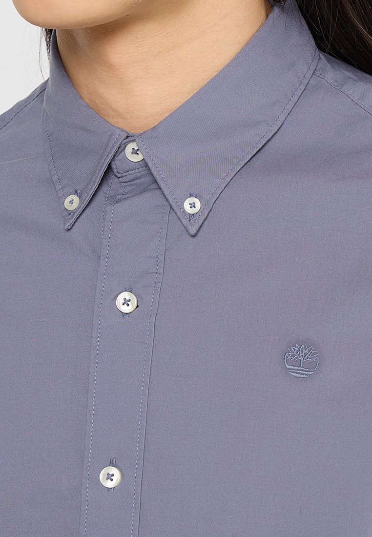 Camisa abotonada gris claro con un cuello clásico, que presenta un pequeño logotipo bordado y botones de plástico redondos. Tejido de algodón suave.