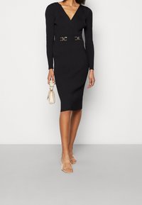 Schwarzes, geripptes Midikleid mit langen Ärmeln, V-Ausschnitt und schmalem Taillenband mit goldfarbener Schnalle. Transparente Riemchenheels und eine kleine metallische Handtasche.