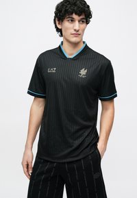 Νεαρός άνδρας που φοράει μαύρο πουκάμισο EA7 με λεπτές ρίγες και τυρκουάζ περιγράμματα, συνδυασμένο με μαύρα σορτς EA7 με επαναλαμβανόμενα λογότυπα.