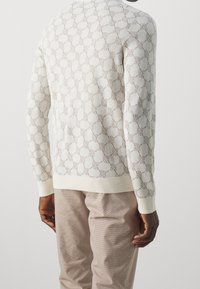 Hellbeiger Strickpullover mit strukturiertem geometrischem Muster. Bündchen und Saum gerippt. Kombiniert mit beigen Texturhosen.