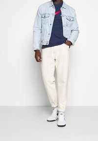 Ljusblå denimjacka över en marinblå t-shirt med röd grafik, i kombination med krämfärgade byxor och vita sneakers med svarta detaljer.