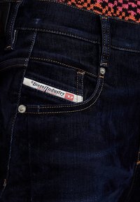Tmavé džíny z denimu s oranžovým prošíváním, s malou kapsou a značkovou etikettou "Diesel Industry Denim Division." Hladký povrch.