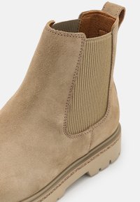 Botín de ante beige hasta el tobillo con paneles laterales elásticos, suela de tracción y tira de pull; presenta una textura suave y costuras reforzadas.