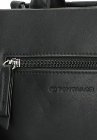 Sac à main en cuir noir avec une poche zippée, présentant des accessoires en métal argenté et le logo "TOM TAILOR" en relief. Texture lisse et design rectangulaire.