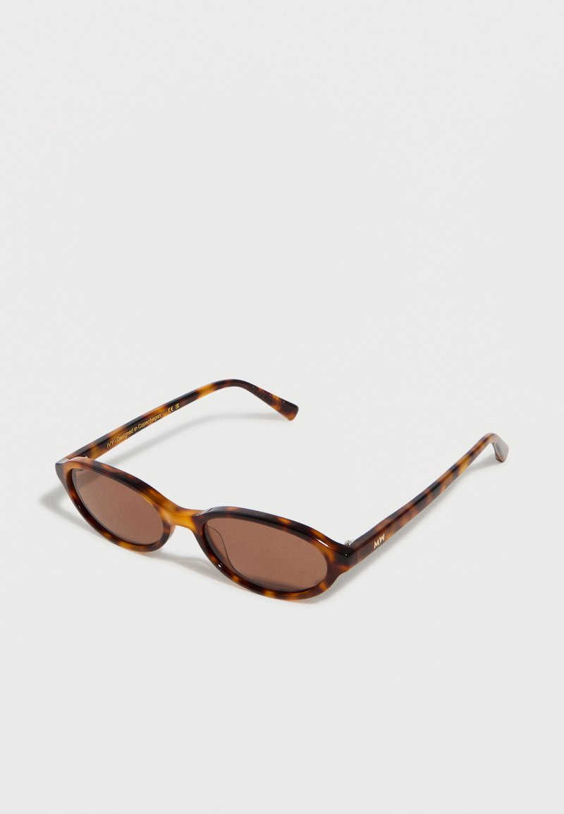 Lunettes de soleil ovales marron avec une monture écaille de tortue et des branches fines reposant sur une surface blanche.