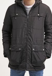 Svart jacka med dragkedja, justerbar huva, två bröstfickor och två nedre fickor. Den har tryckknappsfunktioner och en quiltad textur.