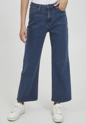 Wijdvallende blauwe spijkerbroek met een hoge taille, voorzien van vijf zakken en contrasterende stiksels. In combinatie met witte sneakers voor contrast.