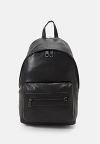 Svart lær ryggsekk med en rund form, utstyrt med et håndtak på toppen, dobbel glidelås-lukking og en glidelåslomme på forsiden. Diskret logo-detalj.