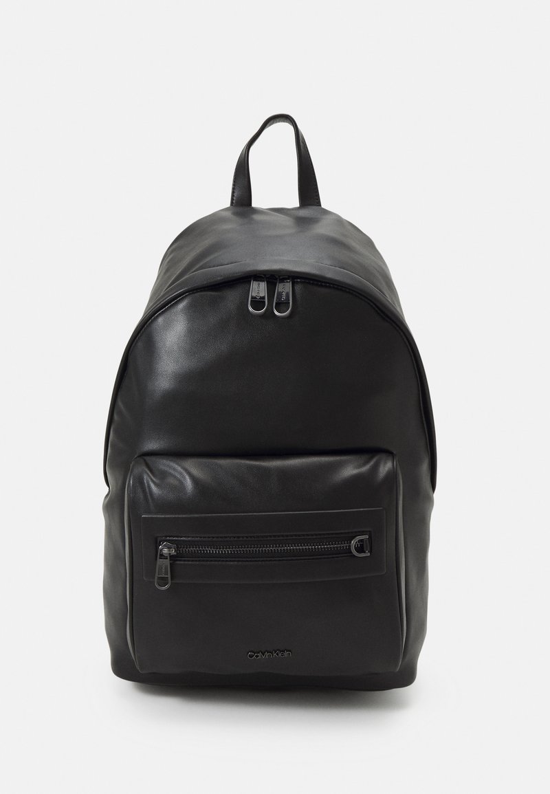 Svart lær ryggsekk med en rund form, utstyrt med et håndtak på toppen, dobbel glidelås-lukking og en glidelåslomme på forsiden. Diskret logo-detalj.