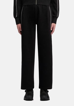 Person trägt schwarze, weit geschnittene Hose mit weißen Seitenstreifen und schwarzen Sneakers, steht vor einem einfarbigen Hintergrund.