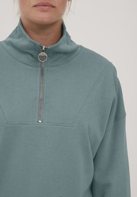 Tealfärgad tröja med hög krage och silverfärgad dragkedja fram. Mjuk tyg, geometrisk design på bröstet och avslappnad passform. Subtila sömnadsdetaljer.