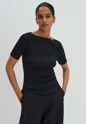 Vrouw draagt een nauwsluitend zwart shirt met korte mouwen en zwarte broek, met haar handen in de zakken, staand tegen een effen lichte achtergrond.