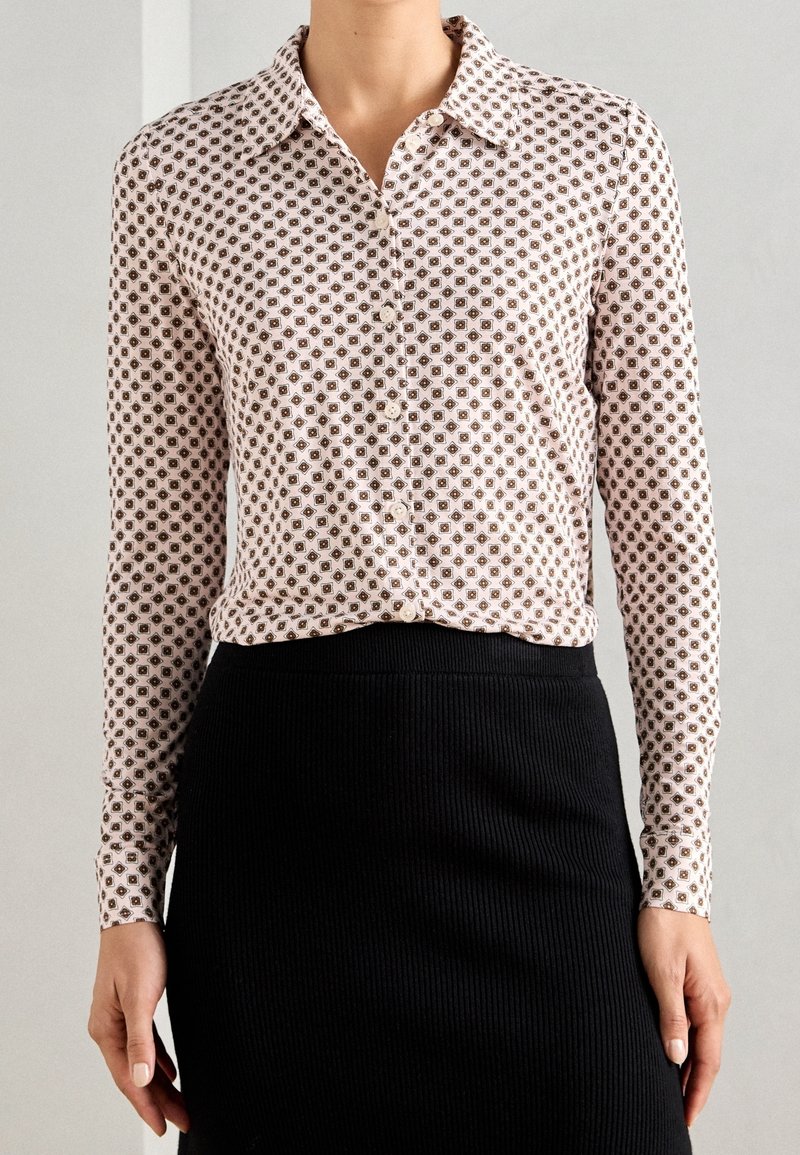 Femme portant un chemisier blanc à manches longues avec un motif géométrique marron et une jupe noire taille haute sur un fond uni.