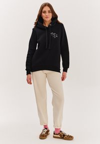 Černá mikina s kapucí a klokanní kapsou, stahovací kapuce a bílým písmovým textem. V kombinaci s béžovými kalhotami a teniskami s leopardím vzorem.