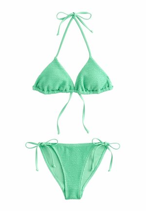 Mujer con bikini verde menta y gafas de sol sosteniendo una bolsa de playa rosa con rayas, de pie en la orilla rocosa con olas del océano al fondo.