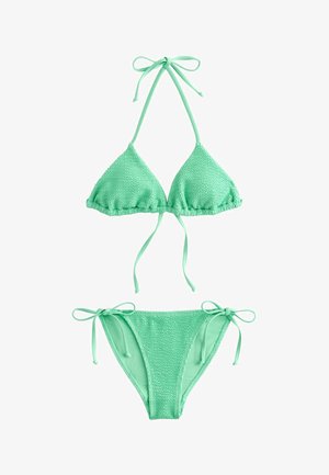 Femeie într-un bikini verde mentă și ochelari de soare ținând o geantă de plajă roz cu dungi, stând pe o țărm stâncos cu valuri oceanice în fundal.
