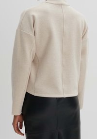 Giacca in misto lana beige con colletto alto, spalle abbassate e taglio dritto. Presenta una cucitura centrale e una texture liscia.