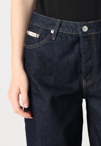 Jeans de mezclilla azul oscuro con corte recto, que presentan una etiqueta de Calvin Klein, botón plateado y costuras en amarillo contrastante.