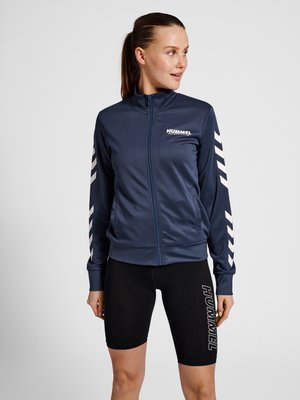 Navy zip-up athletic jacket with white logo and sleeve stripes; paired with black form-fitting shorts featuring white branding. Smooth texture.