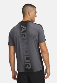 Camiseta atlética gris con acentos negros, que presenta un logo vertical de "ARMOUR" en la parte posterior, mangas cortas y un ajuste aerodinámico.