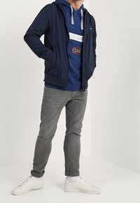 Sudadera azul con cremallera y detalles grises, usada debajo de una chaqueta azul marino, jeans grises y zapatillas blancas. El conjunto incluye un gráfico de bandera en la sudadera.