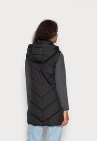 Schwarze Steppweste mit Kapuze und Zickzack-Quiltmuster, kombiniert mit einem grauen Langarmshirt und hellblauen Hosen.