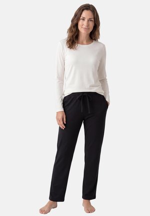 Femme aux cheveux bruns ondulés portant une chemise blanche à manches longues et un pantalon noir à cordon, debout pieds nus sur un fond blanc.