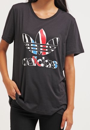 Vrouw draagt een zwart Adidas T-shirt met een rood-wit-blauw gestreept trefoil-logo en bijpassende tekst aan de voorkant.