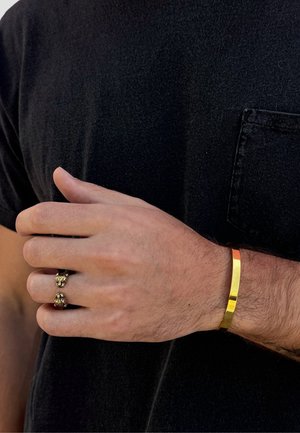 Mandshånd iført et guldarmbånd og en åben guldring med dyrehoveddesigns, mod en sort kortærmet skjorte med brystlomme.