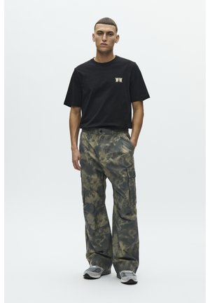 Jaunas vyras stovintis, dėvintis juodą marškinėlį su logotipu ir laisvus kamufliažo cargo kelnes bei pilkus sportbačius, priešais paprastą šviesią foną.