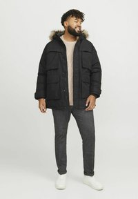 Parka acolchada negra con capucha forrada de piel, que cuenta con múltiples bolsillos en la parte frontal. Lleva puesta una ligera sudadera de punto beige claro y pantalones grises.