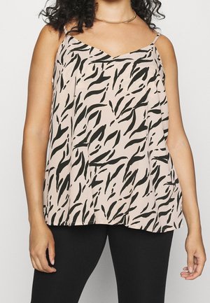 Mujer con una blusa sin mangas beige con rayas abstractas negras similares a las de una cebra y pantalones negros, de pie frente a un fondo liso.