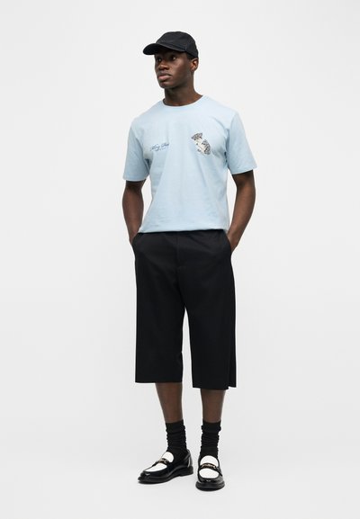 Άνδρας που φοράει μαύρο καπέλο, ανοιχτό μπλε t-shirt με σχέδιο, μαύρα σορτς μέχρι το γόνατο, μαύρες κάλτσες και μαύρα και λευκά μοκασίνια, στέκεται με τα χέρια στις τσέπες.