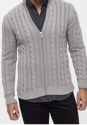 Homme portant un cardigan zippé gris clair à côtes torsadées par-dessus une chemise boutonnée gris charbon foncé et un pantalon noir.