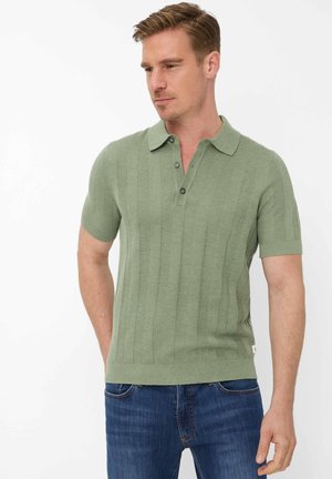 Man die een lichtgroen poloshirt met korte mouwen en een verticale breipatroon draagt, met een blauwe spijkerbroek, staand met handen nabij de zakken, naar beneden kijkend.