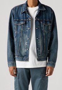 Man som bär en blå denimjacka över en vit t-shirt och grå cargobyxor, stående mot en enfärgad ljus bakgrund.