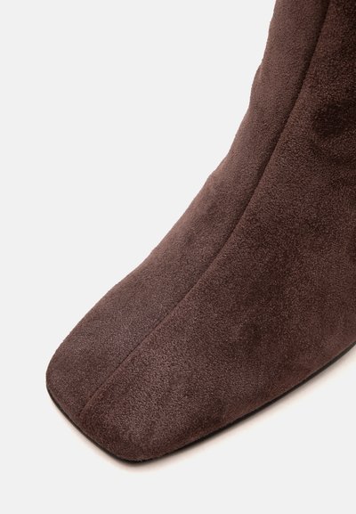 Gros plan d'une botte en daim marron avec un bout carré et un détail de couture centrale visible sur un fond uni.