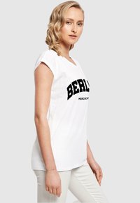 Hvid bomulds T-shirt med korte opkrøllede ærmer, der har en fed sort "BERLIN" tekst og en mindre "MERCHANDISE" nedenunder.